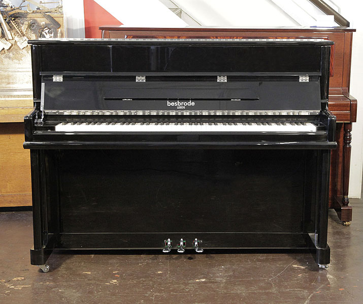 A brand new, Besbrode SU112 upright piano with a black case and chrome fittings. Piano features a slow fall mechanism, has an eighty-eight note keyboard and three pedals.
