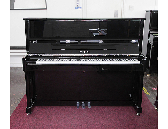 A brand new, Feurich Model 122 upright piano with a black case and chrome fittings. Piano has an eighty-eight note keyboard and three pedals.